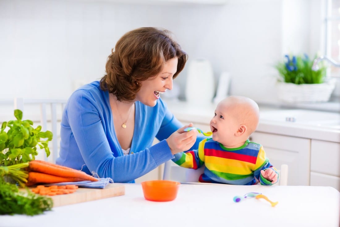 una giovane madre che nutre il suo bambino con un cucchiaio che è seduto nel suo seggiolone