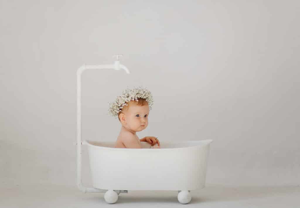foto da un servizio fotografico per bambini. un bambino con una corona di fiori bianchi in testa, è seduto in una vasca da bagno tutta bianca con ruote. ha un rubinetto bianco sopra la testa