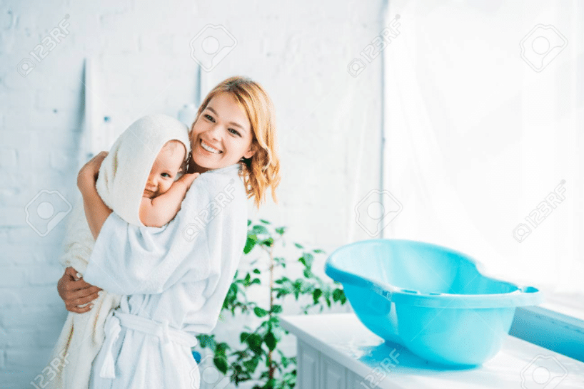 una madre in accappatoio bianco porta un bambino nel suo asciugamano beige che è appena uscito dalla sua vasca da bagno blu