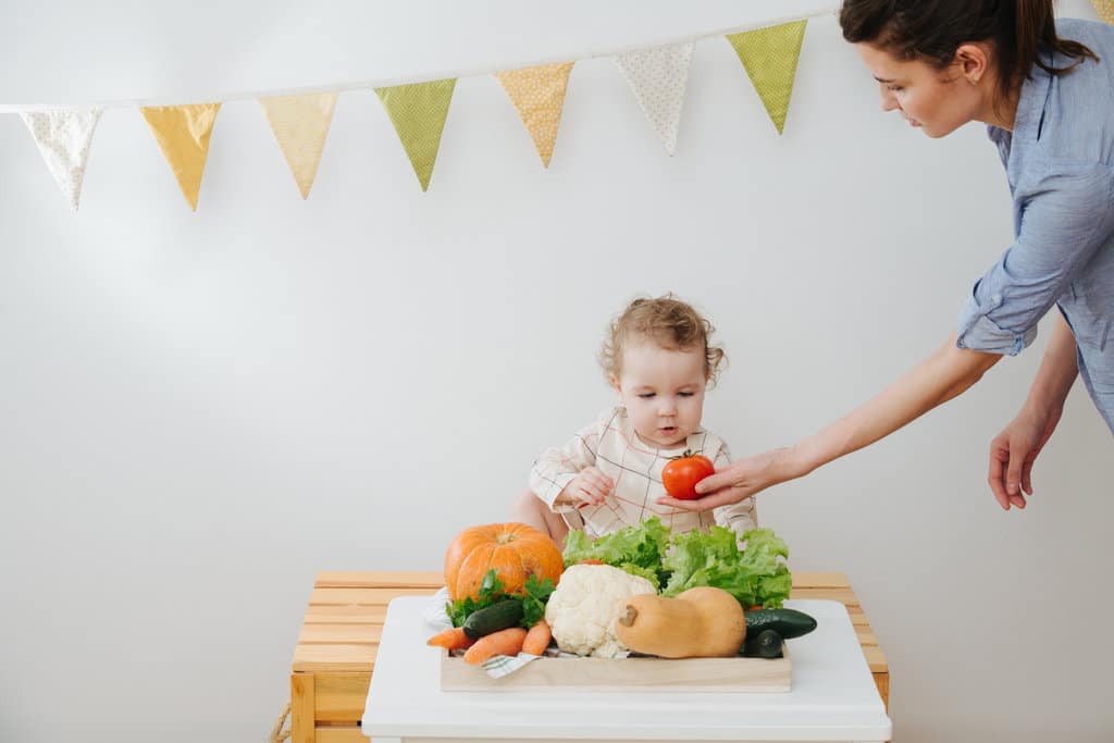 Un bambino sta davanti a un tavolo con verdure fresche e guarda con stupore un pomodoro che la madre gli mette davanti al viso