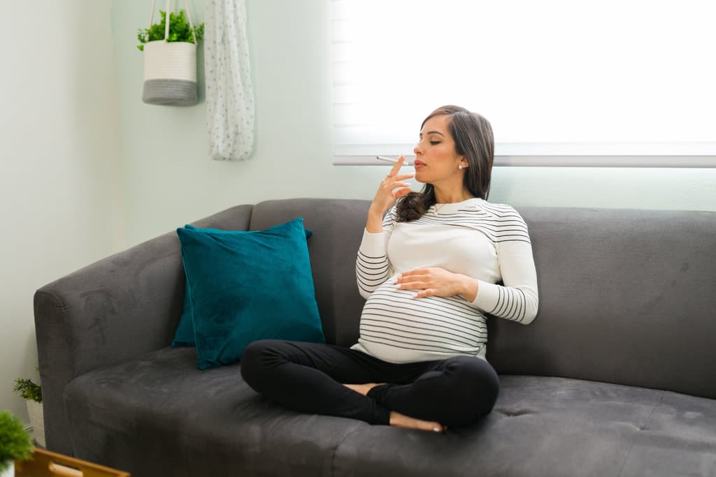 una donna incinta fuma una sigaretta, seduta a gambe incrociate su un divano grigio e corre il rischio di avere un bambino che tossisce