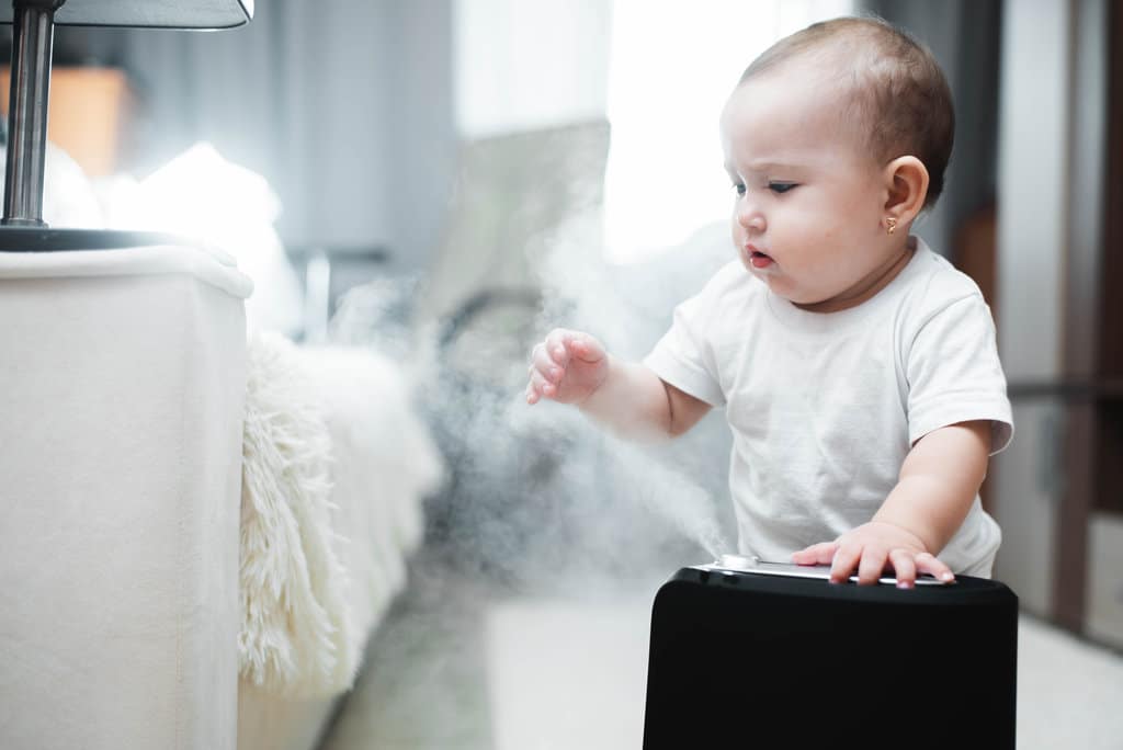 un bambino in un body bianco a maniche corte gioca con un umidificatore a vapore in una stanza accanto a un letto